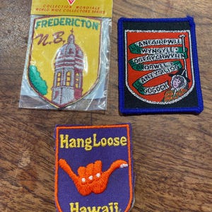 May include: Three embroidered souvenir patches. One is in a plastic sleeve labelled Voyageur, featuring a yellow shield with a building and the text Fredericton N.B. Another is a rectangular patch with green signs and a person. One is purple with an orange hand sign and the text Hang Loose Hawaii.