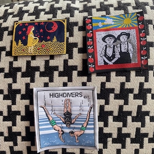 May include: Three embroidered patches with various designs. One shows a woman in a yellow dress under a red moon and stars. Another depicts two figures in black and white with a red border. The third patch says "HIGH DIVERS" and shows divers.