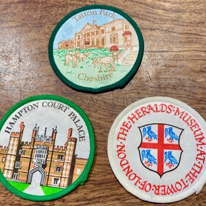 May include: Three circular embroidered patches featuring illustrations of Tatton Park in Cheshire with deer, Hampton Court Palace, and the Heralds' Museum at the Tower of London with a red cross and blue bird crest on a white background.
