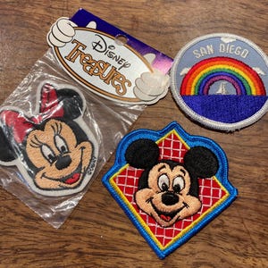May include: Three embroidered patches featuring Disney characters. One patch shows Minnie Mouse in a plastic bag with a Disney Treasures header card. Another is a circular San Diego patch with a rainbow. The third is a diamond-shaped Mickey Mouse patch.