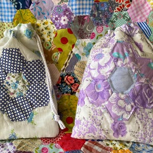May include: Two fabric drawstring pouches featuring quilted hexagon flower motifs. The left pouch is white with a blue polka dot and floral flower. The right pouch is white with a purple floral flower. Both have white braided rope drawstrings.