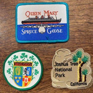 May include: Three embroidered patches: a rectangular patch featuring the Queen Mary and Spruce Goose with text, a circular patch displaying the Ireland coat of arms with shamrocks, and an irregular tan patch depicting a Joshua Tree National Park scene.