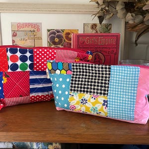May include: Two quilted zip pouches featuring a patchwork design of various fabric patterns including polka dots, gingham, floral prints, and houndstooth in vibrant colours like red, blue, pink, yellow, and black with matching pink zips.