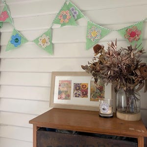 May include: A fabric bunting featuring green striped triangular flags with colourful floral appliqués, hung above a wooden cabinet. The cabinet holds a framed botanical print, a glass vase with dried flowers, and a small candle in a glass jar.