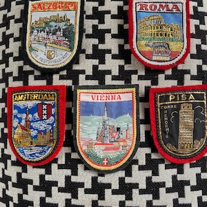 May include: Embroidered travel patches showcasing famous landmarks. Patches include "Salzburg," "Roma," "Amsterdam," "Vienna," and "Pisa." Each patch has a detailed design and a red border, set against a black and white houndstooth pattern.