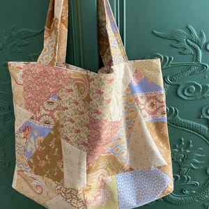 May include: A patchwork tote bag constructed from various fabric scraps featuring floral, paisley, and geometric patterns in shades of pink, yellow, blue, and cream. The fabric includes the printed text "go am of me" on one of the light-coloured panels.
