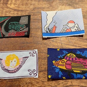 May include: Four embroidered patches with different designs. One shows a green car, another a person in the water, an angel, and a rocket ship. The patches are rectangular and have a woven texture.