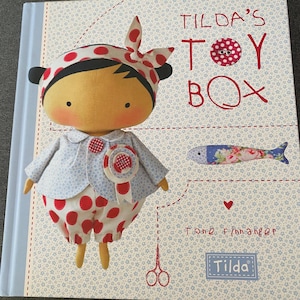 May include: The cover of "Tilda's Toy Box" by Tone Finnanger features a fabric doll in a red and white polka dot outfit and headscarf. The book has a light blue floral patterned background, and a fabric fish illustration.
