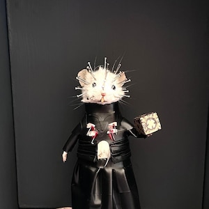 May include: A taxidermy white mouse figurine wearing a black outfit with a high collar and a long skirt. The mouse has silver pins inserted into its head and chest, and it holds a small, patterned cube in one paw. A long tail extends outward.