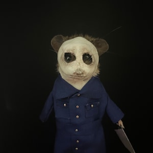 May include: A taxidermy mouse figure wearing a blue collared jumpsuit with silver buttons. The mouse has a white, textured mask covering its face with dark, hollowed-out eye sockets and holds a small, silver-colored blade in its left hand.