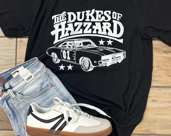 May include: Black t-shirt with a white graphic of a car and the text "The Dukes of Hazzard". The shirt is paired with light-wash denim shorts and white sneakers with black accents and a brown sole.