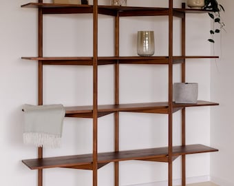 May include: A dark brown wooden shelving unit with several shelves, displaying decorative items including a framed abstract art piece, a glass vase with dried plants, a potted plant, a grey pot, and a light green throw.