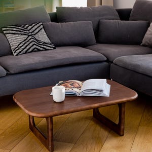 Peut inclure: Une table basse en bois brun foncé avec un plateau rectangulaire arrondi et des pieds inclinés. Une tasse blanche et un livre ouvert reposent sur la table. Un canapé d'angle gris et un coussin à motifs sont en arrière-plan.