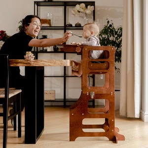 May include: A wooden toddler step stool in a warm brown finish. The step stool has a built-in safety rail and is designed to bring a child up to counter height. A person is feeding a baby in the step stool. The scene takes place in a well-lit dining room.