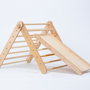 May include: A wooden climbing triangle with a slide attached. The triangle has a ladder on one side and the slide is on the other side. The triangle is made of natural wood and has a light brown color.