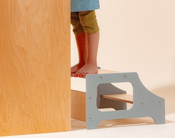 Puede incluir: Un taburete de madera de dos peldaños con un marco pintado de gris y peldaños de madera natural. El taburete está diseñado para que los niños puedan alcanzar superficies más altas.