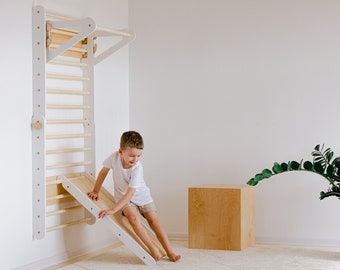 May include: A white wooden climbing wall with a slide attached. A young child is sliding down the slide. The wall has a ladder and a monkey bar. The slide is made of wood and has a white finish.