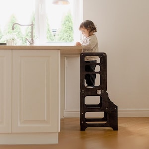 May include: A dark brown wooden kitchen step stool. The step stool has a rectangular frame with cut-out sections. A child is standing on the stool near a light-coloured countertop and a window. The stool is designed to help children reach higher surfaces.