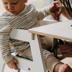 May include: A white high chair with a wooden tray. A baby is sitting in the chair and reaching for the tray.