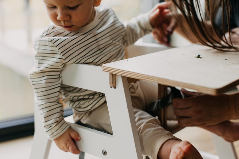 May include: A white high chair with a wooden tray. A baby is sitting in the chair and reaching for the tray.