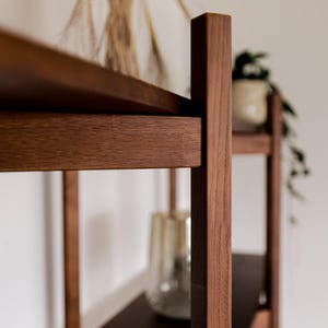 May include: A close-up of a wooden bookshelf with dark brown shelves and vertical supports. The wood has a natural grain and a warm, reddish-brown hue. A glass vase and a potted plant are visible on the shelves, adding a touch of decor.