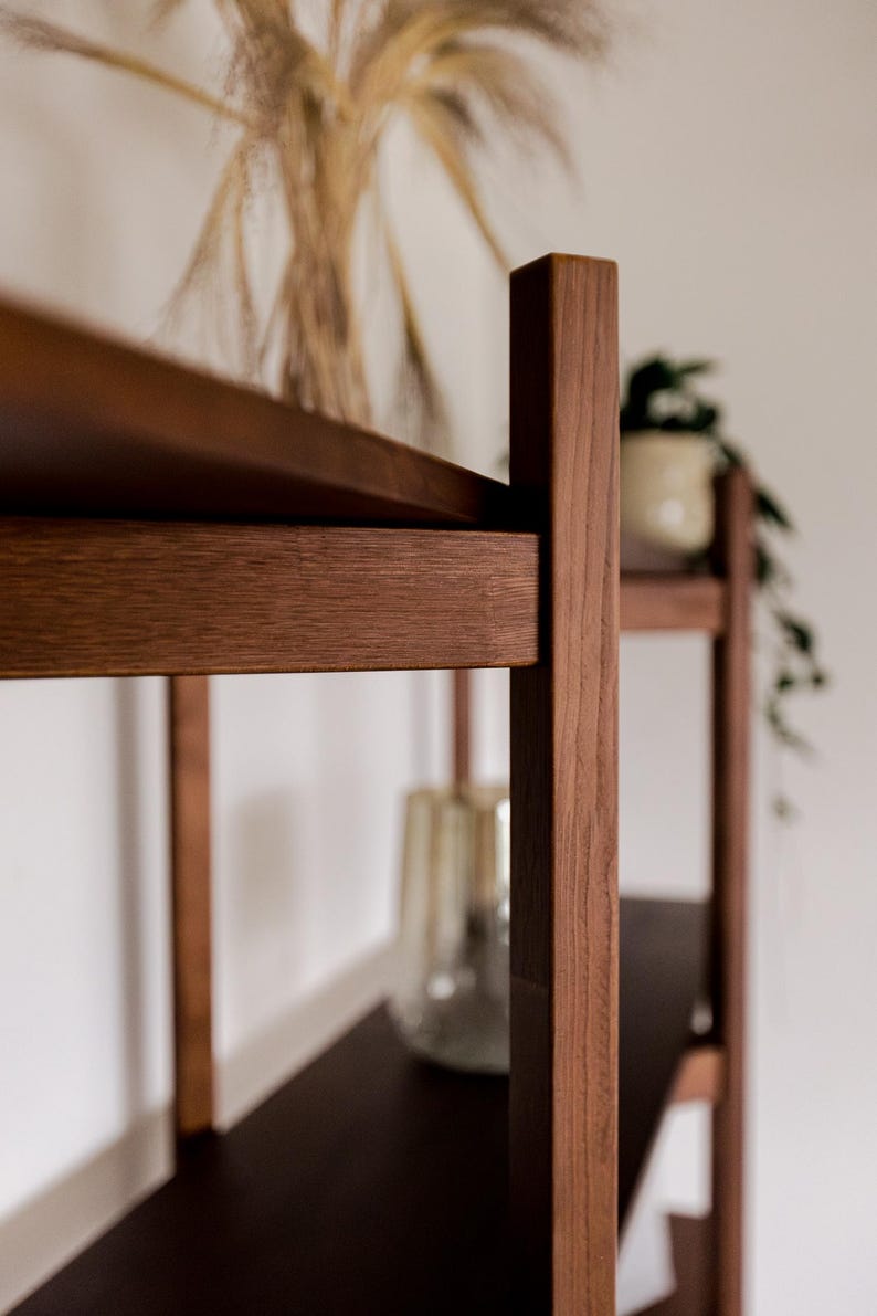 May include: A close-up of a wooden bookshelf with dark brown shelves and vertical supports. The wood has a natural grain and a warm, reddish-brown hue. A glass vase and a potted plant are visible on the shelves, adding a touch of decor.
