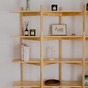 May include: A light-colored wooden bookshelf with multiple shelves. The shelves display various decorative items, including books, candles, a framed picture, a small statue, and a woven tray. Tall grass in a clear vase adds a natural element.