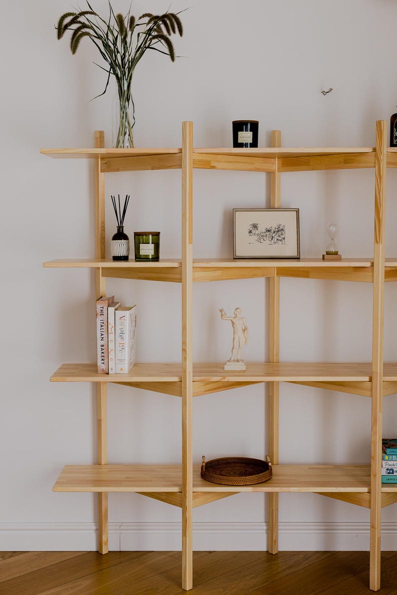 May include: A light-colored wooden bookshelf with multiple shelves. The shelves display various decorative items, including books, candles, a framed picture, a small statue, and a woven tray. Tall grass in a clear vase adds a natural element.