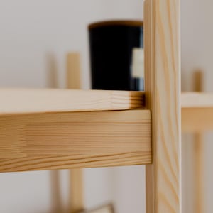 May include: Close-up of a light-colored wooden shelf with a black container on the top shelf. The shelf has a simple, minimalist design with visible wood grain. The background is a plain white wall.