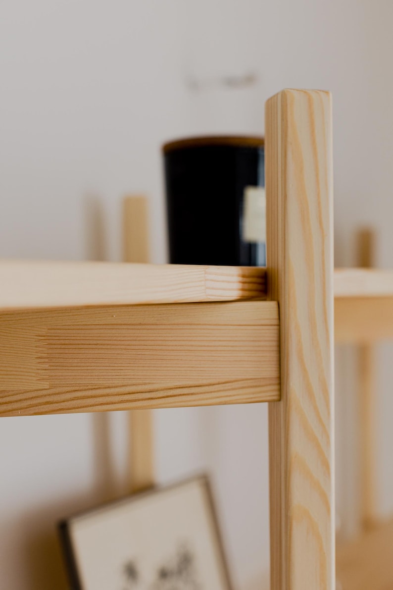 May include: Close-up of a light-colored wooden shelf with a black container on the top shelf. The shelf has a simple, minimalist design with visible wood grain. The background is a plain white wall.