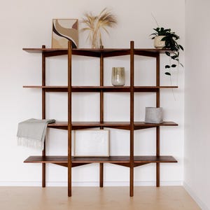 May include: A dark wood, mid-century modern style shelving unit with four shelves. The shelves hold decorative items like a framed print, a vase with dried plants, a potted plant, and a small gray container. A light gray blanket is draped on one shelf.
