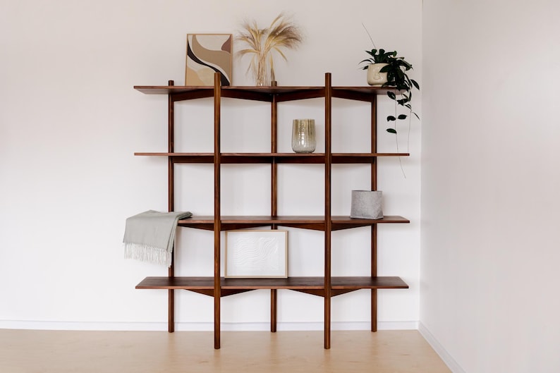 May include: A dark wood, mid-century modern style shelving unit with four shelves. The shelves hold decorative items like a framed print, a vase with dried plants, a potted plant, and a small gray container. A light gray blanket is draped on one shelf.