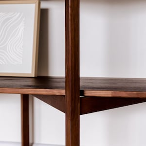 May include: A dark brown wooden shelf unit with a framed artwork. The shelves are supported by vertical and angled wooden supports. The artwork has a light-colored abstract design. The unit is against a white wall.