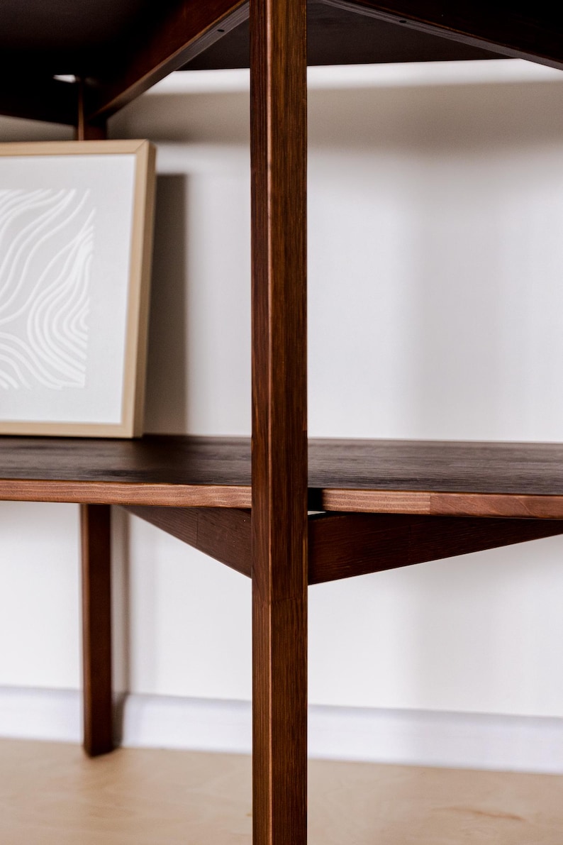 May include: A dark brown wooden shelf unit with a framed artwork. The shelves are supported by vertical and angled wooden supports. The artwork has a light-colored abstract design. The unit is against a white wall.