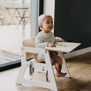 May include: A white wooden high chair with a light wood tray and a gray strap. A young child is sitting in the chair, wearing a striped shirt and white pants.