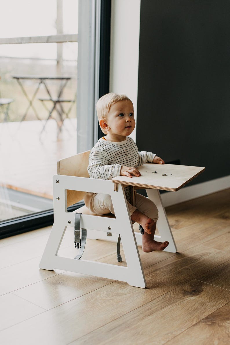 May include: A white wooden high chair with a light wood tray and a gray strap. A young child is sitting in the chair, wearing a striped shirt and white pants.