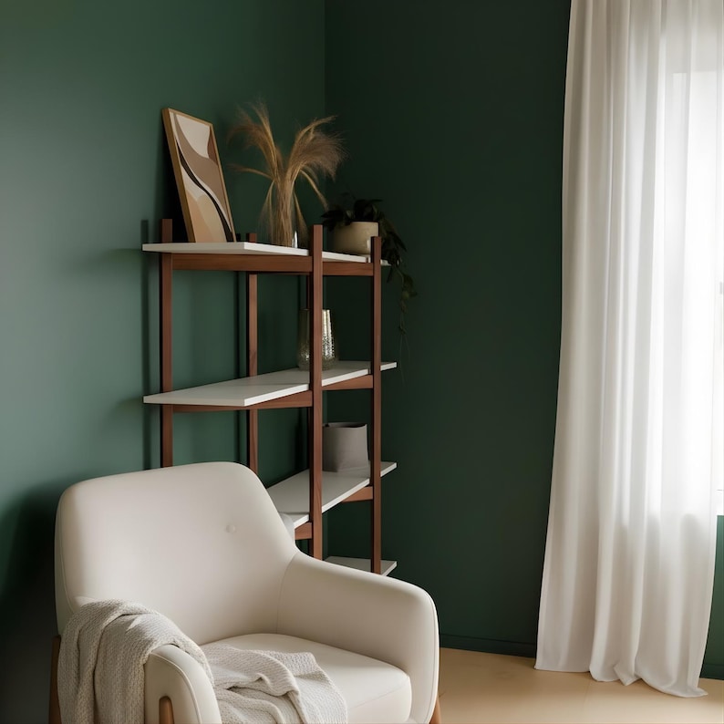 May include: A modern living room corner with a white armchair and a wooden bookshelf. The bookshelf has white shelves and brown supports, displaying decorative items. A framed artwork and plants add to the room's aesthetic. The walls are painted a deep green, and a white curtain hangs in the window.