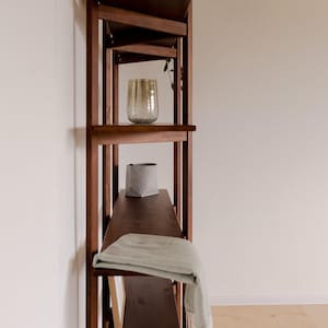 May include: A dark brown wooden bookshelf with angled shelves. The shelves hold a decorative glass, a gray felt basket, and a folded light green blanket. Dried plants are placed on the top shelf. The bookshelf is against a white wall.