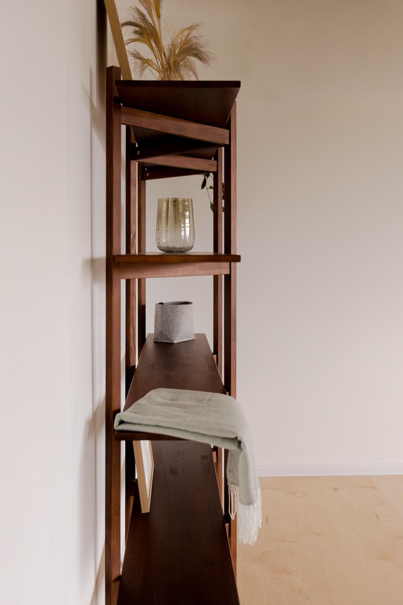 May include: A dark brown wooden bookshelf with angled shelves. The shelves hold a decorative glass, a gray felt basket, and a folded light green blanket. Dried plants are placed on the top shelf. The bookshelf is against a white wall.
