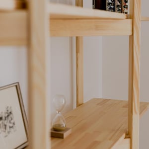 May include: A light-colored wooden shelf unit with three shelves. The top shelf holds several brown bottles with cork stoppers. An hourglass and framed artwork are on the lower shelves. The unit is made of light-colored wood.