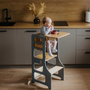 May include: A gray and natural wood toddler kitchen helper tower with a small table. A baby is sitting on the tower, eating from a red bowl. The tower has a step and a platform for the child to stand on.