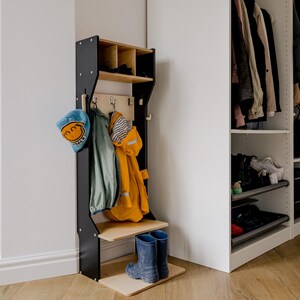 May include: A black wooden coat rack with a shelf and hooks, holding a blue hat, green jacket, and yellow raincoat. Two blue rain boots stand on the base. A white closet is in the background.