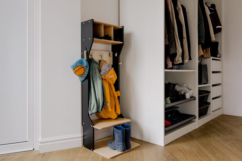 May include: A black wooden coat rack with a shelf and hooks, holding a blue hat, green jacket, and yellow raincoat. Two blue rain boots stand on the base. A white closet is in the background.