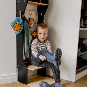 May include: A black and wood entryway organizer with a shelf, hooks, and cubbies. A child is sitting on the shelf, wearing blue boots. A blue hat and jacket hang on the organizer. The organizer is against a white wall.