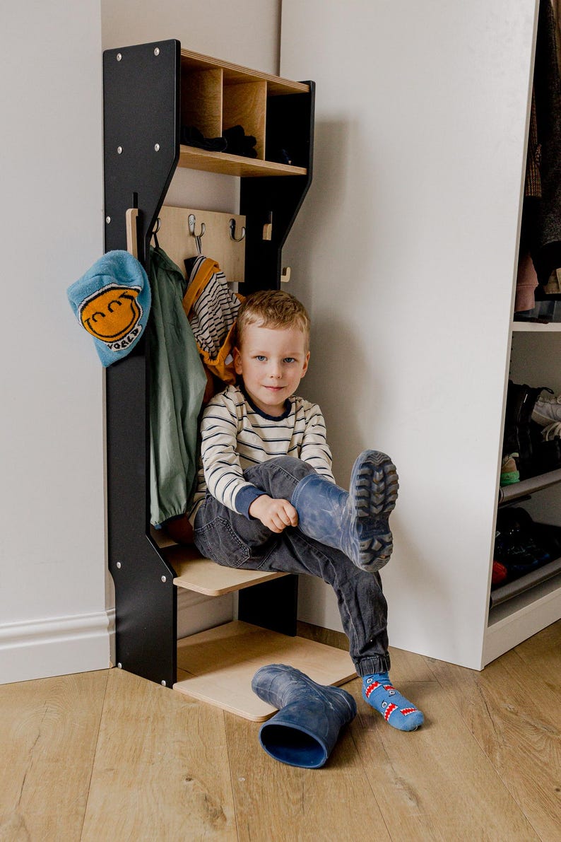 May include: A black and wood entryway organizer with a shelf, hooks, and cubbies. A child is sitting on the shelf, wearing blue boots. A blue hat and jacket hang on the organizer. The organizer is against a white wall.