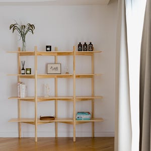 May include: A light-colored wooden shelving unit with multiple shelves, displaying various decorative items. The shelves hold books, framed art, and small bottles. The unit is against a white wall, with a glimpse of a window and curtains on the right.