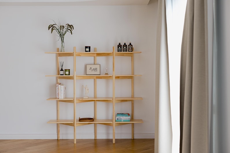 May include: A light-colored wooden shelving unit with multiple shelves, displaying various decorative items. The shelves hold books, framed art, and small bottles. The unit is against a white wall, with a glimpse of a window and curtains on the right.