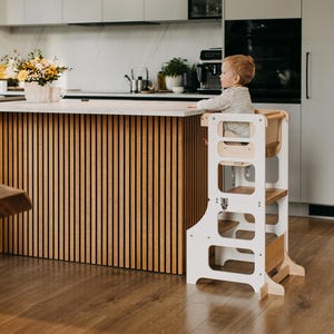 May include: A white wooden learning tower with a child sitting on the top step. The tower has three steps and a safety rail. The tower is in a kitchen with a wooden countertop and a light wood floor.