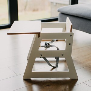 May include: White wooden high chair with a removable tray and a gray five-point harness. The chair is on a wood floor.