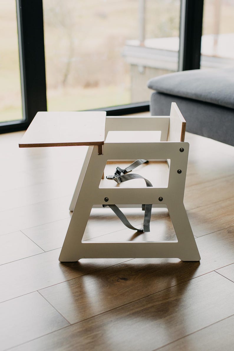 May include: White wooden high chair with a removable tray and a gray five-point harness. The chair is on a wood floor.